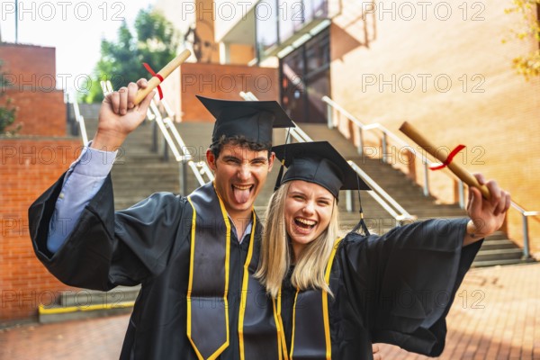 Young man and woman friends wearing graduation caps and gowns, cheering and holding diplomas outside a university campus, celebrating academic achievement and a new journey