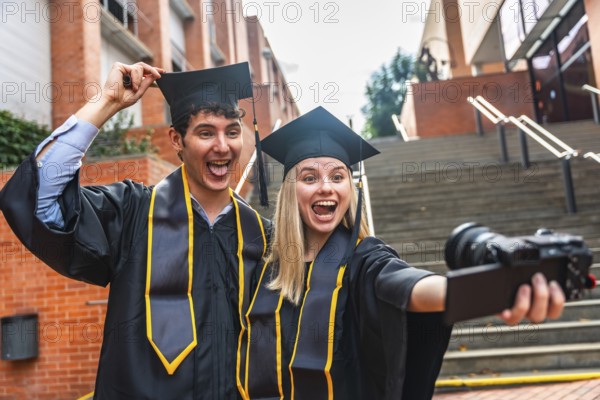 Two cheerful university graduates, a man and a woman, wearing cap and gown, smiling and making funny faces while holding a camera for a selfie on their graduation day