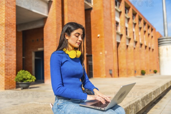 Young woman in casual clothing engaging in remote learning or project work on a laptop, sitting outside on a university campus building in daylight with headphones around her neck