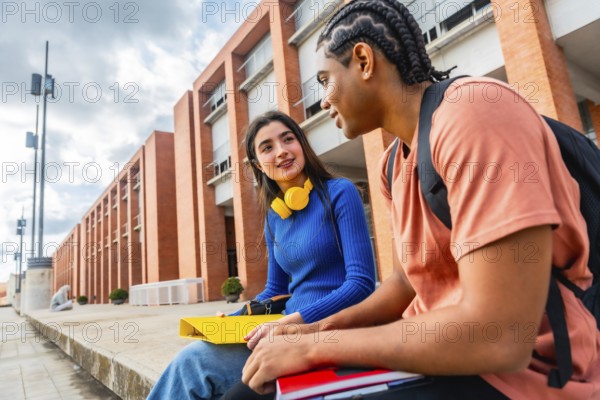 Two diverse young students engaging in a friendly conversation while sitting on a ledge on the university campus, holding notebooks and textbooks, representing education and friendship