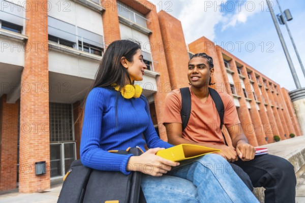 Two young diverse students are sitting on a wall outside a university building, engaging in a friendly conversation, holding school supplies, and representing modern education and campus life