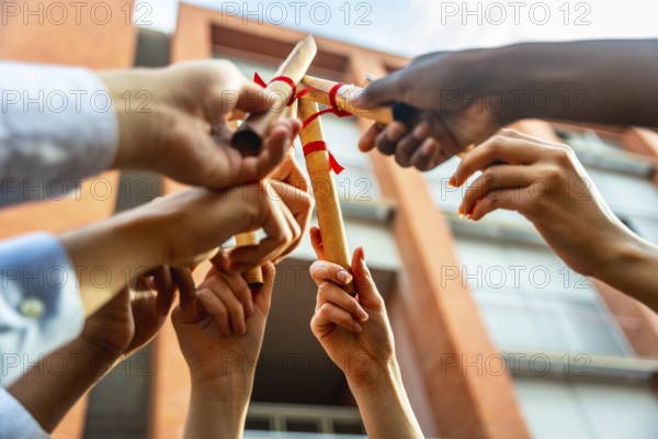 Diverse group of university students holding rolled up diplomas with red ribbons in the air against a campus building, celebrating their academic achievement and future success