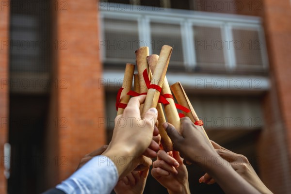 Graduates from various backgrounds raise their rolled up diplomas, tied with red ribbons, celebrating their educational achievement and success at the university