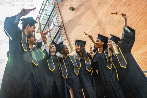 Diverse group of joyful young graduates in black caps and gowns celebrating on campus, holding diplomas and smiling together after completing their university degrees