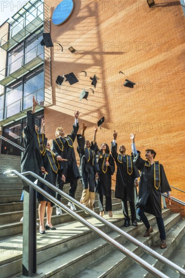 Diverse group of young adult university graduates celebrating a successful academic achievement by tossing their graduation caps into the air on stairs in front of a modern brick building