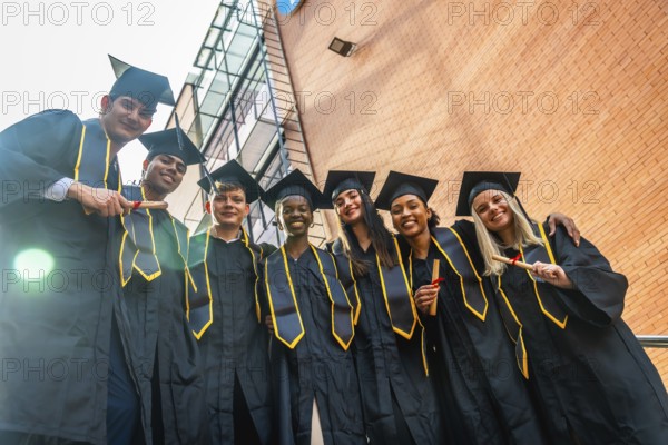 Diverse group of joyful university graduates in caps and gowns standing outdoors by campus buildings, proudly holding diplomas and celebrating their academic achievement and new beginnings