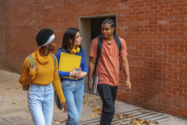 Three diverse young university students walking along a brick wall, carrying backpacks and books, engaging in conversation while heading to class on an autumn day