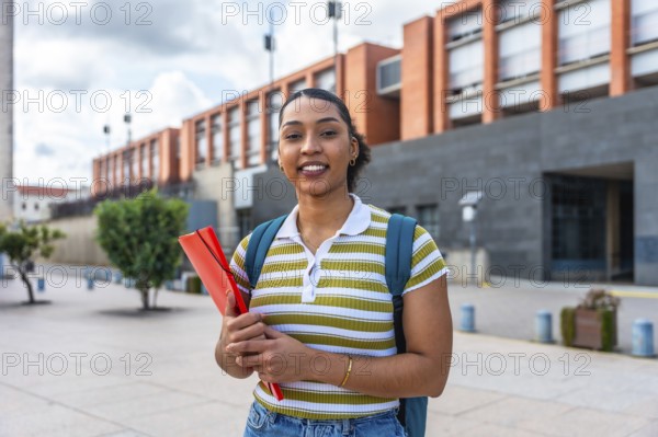 Young female student smiling while holding a folder and wearing a backpack, standing outdoors on a university campus with modern buildings in the background