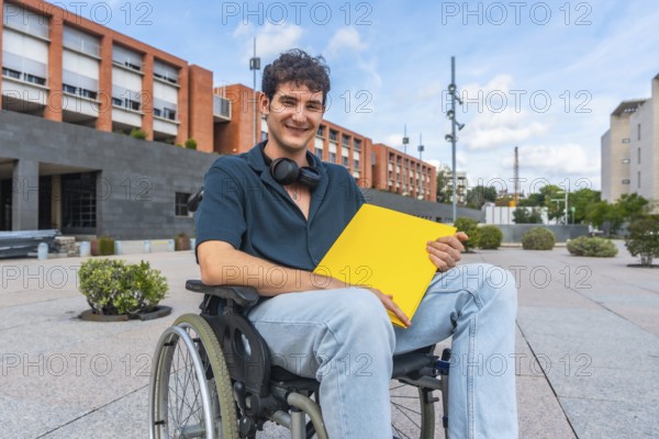 Young male student in a wheelchair smiling, holding a yellow folder, and wearing headphones around his neck, exploring the accessible university campus on a sunny day