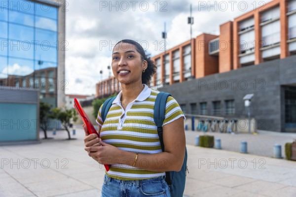 Young latina student smiling and walking on a university campus while carrying a backpack and a red folder, representing education, youth, and future success