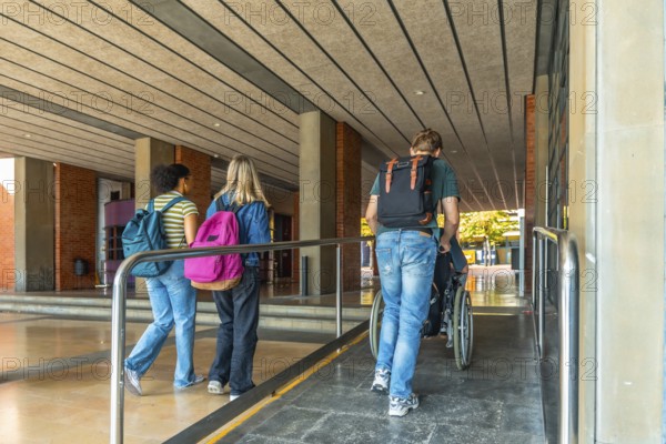 Diverse students entering a university building, one student helping another in a wheelchair using an accessible ramp, symbolizing inclusion and support in education