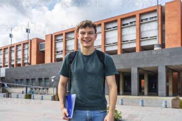 Young male student with backpack and folders standing outdoors on campus, smiling confidently at camera, symbolizing higher education, ambition, and fresh beginnings