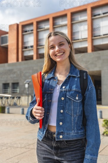 Happy young blonde woman with a backpack and a red folder, smiling confidently while standing outside a modern university building on a sunny day, representing education and successful student life