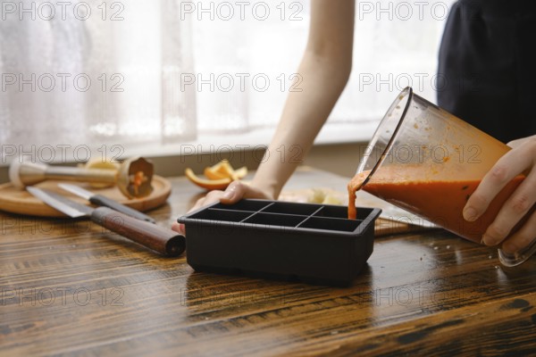 A person is pouring a blend of sea buckthorn, orange and ginger into a silicone ice tray on a wooden table in a kitchen. Various kitchen tools and fruit are visible. The scene shows food preparation in the morning light