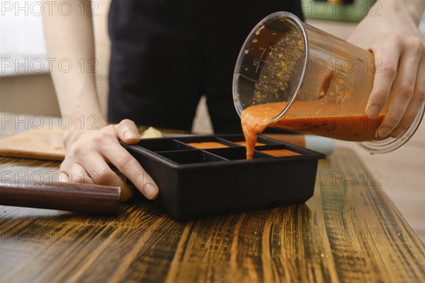 A person pours a blend of orange, sea buckthorn and ginger from blender container into a silicone ice cube tray with multiple sections. The setting is a kitchen with kitchen tools nearby. The time of day is not clear