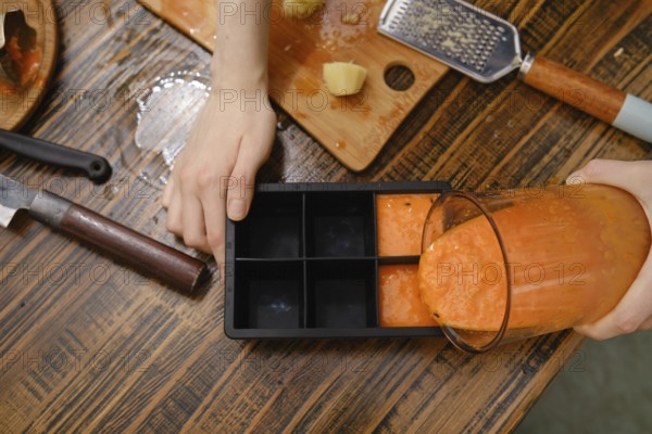 A person is pouring fruit mixture from a blender into a silicone ice cube tray. The table has ginger, a knife, and a grater. This scene shows cooking and meal prep in the kitchen