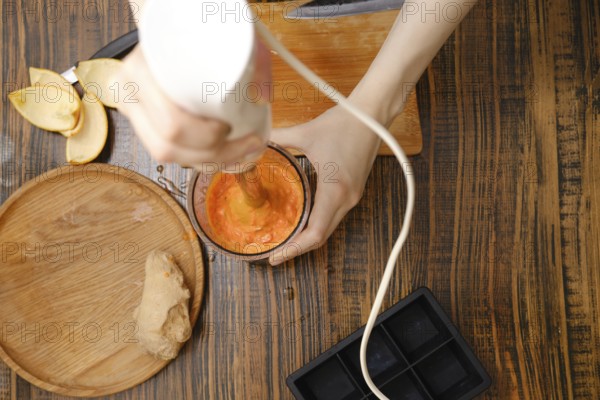 A person uses a hand blender to mix fruits and ginger in a cup. The setting looks like a kitchen during daytime with natural light