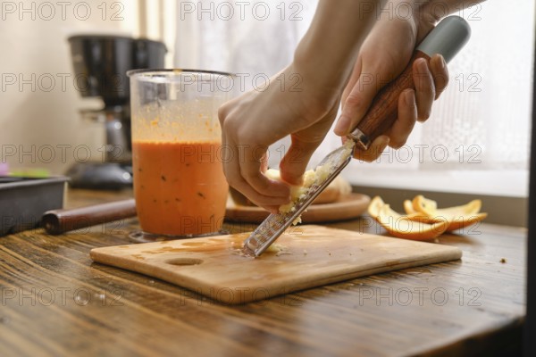 A person is grating fresh ginger on a wooden cutting board in a kitchen. There is a blender with orange liquid nearby. Fresh apple slices rest on the side