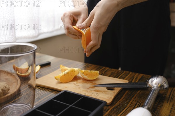 A person peels orange slices on a wooden cutting board in the kitchen during the process of preparation of sea buckthorn and ginger beverage