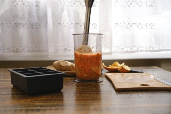 Mixing sea buckthorn and orange in a jar with immersion blender on a wooden table near the window . Sunlight comes in through the window and brightens the space