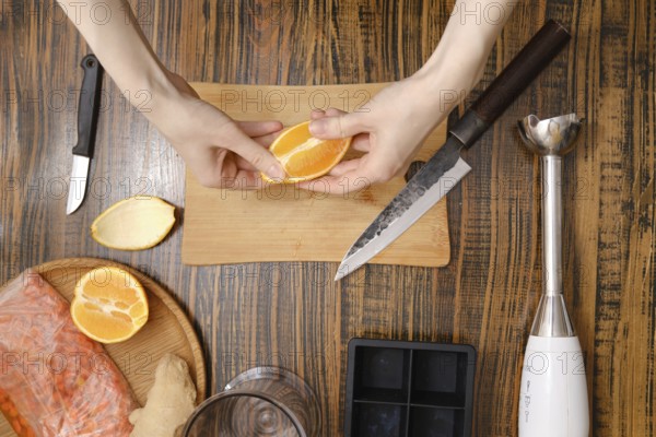 Hands peel an orange over a cutting board. Knives, a blender, and other items surround. The kitchen is busy with various fresh ingredients ready for making frozen ice cubes if vitamin beverahe
