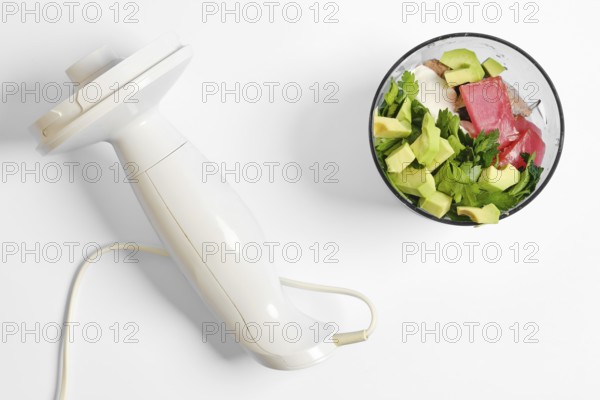A white hand blender rests beside a bowl filled with chopped tuna, avocados, and herbs. The process of cooking fish paste