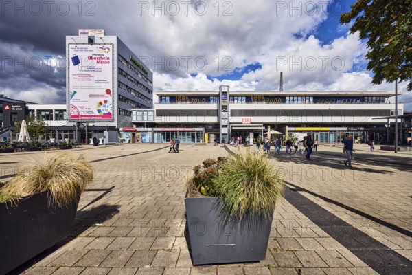 Train station, modern architecture, apartment building, concrete paving stone walkway, pedestrian as accessories, plant pot, rainy mood, blue sky, cumulus congestus clouds, cumulus congestus clouds, Am Bahnhofsplatz, Kehl, Upper Rhine Plain, Ortenau district, Baden-Württemberg, Germany