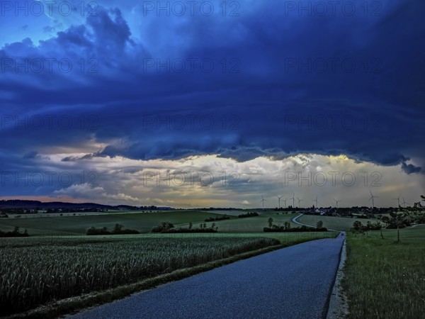 Supercell, approaching thunderstorm with impressive and threatening cloud formation, a thunderstorm over a country road and a village with wind turbines in the background, Baden-Württemberg, Germany
