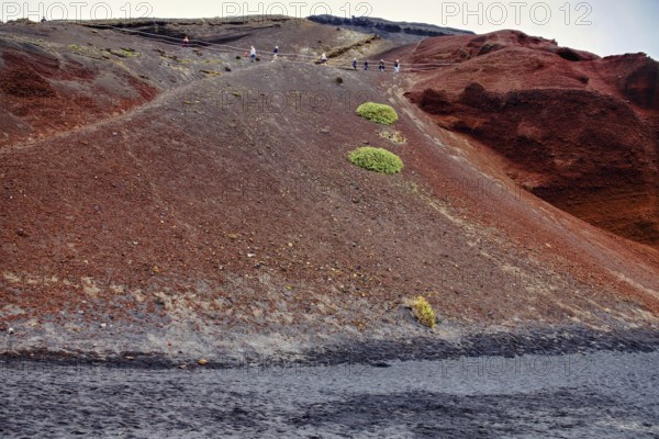 Red volcanic landscape with sparse vegetation and people at the top of a hill, El Golfo Lanzarote