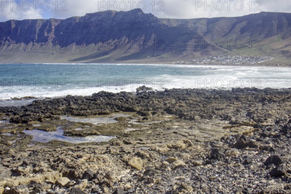 Rough coastal landscape with rocks in the foreground and mountains in the background (Risco de Famara) under a cloudy sky, Playa Caleta de Famara, Teguise Lanzarote