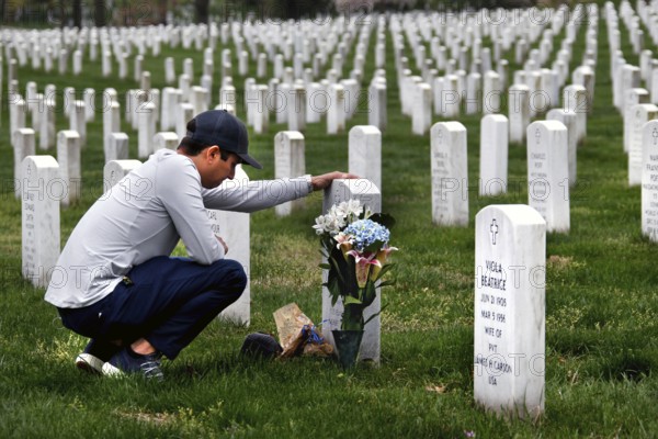 A man kneels in front of a tombstone in a large cemetery and deposits a bouquet of flowers, Washington D.C, USA