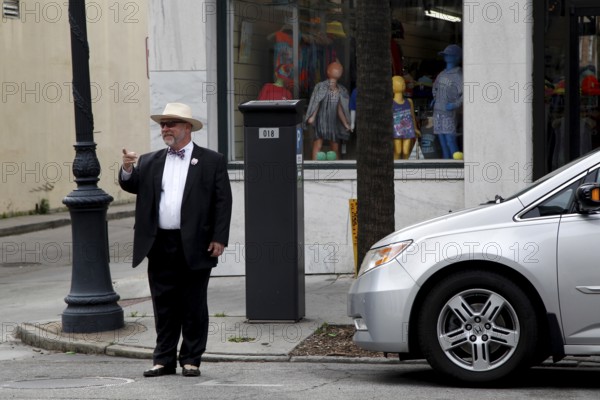 Elderly man in formal clothes stands in front of a shop window on a street corner. Modern car parked next to it, Savannah, Georgia, USA