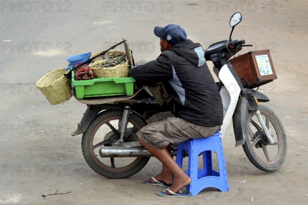 Dealer pauses on a blue stool next to his motorbike loaded with baskets, Oulidia, Doukkala-Abda, Morocco