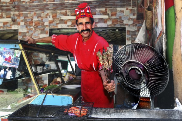 Smiling chef in red outfit offers brochettes at a barbecue area in Moulay Idris, Moulay Idris, null, Morocco