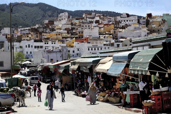 Bustling marketplace in a mountainous landscape with numerous stalls and people in the foreground, Moulay Idris, Fès-Meknès, Morocco