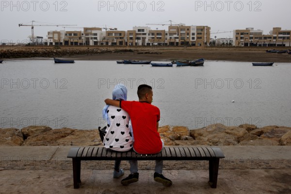 A couple sits on a bench on the riverbank with a view of modern buildings and cranes, Rabat, Salé, Morocco