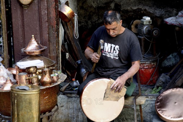 Craftsman repairs copper dishes surrounded by traditional tools and metalwork, Fez, Fez el Bali, Morocco
