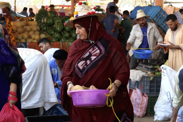 Woman wearing a headscarf carrying a basket full of vegetables at a busy market, Guelmim, Morocco