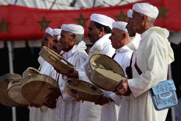 Men wearing traditional clothes holding drums in front of a backdrop with Berber decoration, El Kelâa m'Gouna
