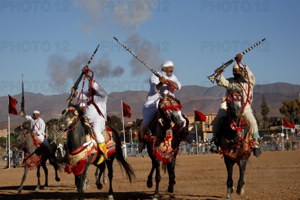 Three horsemen pass each other with rifles in traditional clothing, surrounded by dusty landscape, El Kelâa m'Gouna, Morocco