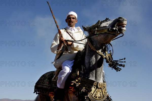 A man in traditional traditional costume rides a magnificently decorated horse under a blue sky, El Kelâa m'Gouna, Morocco