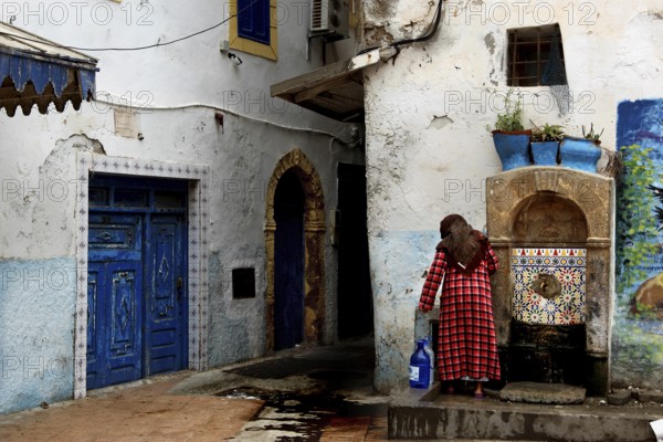 A woman draws water from a spring next to a blue gate in the old town, Essaouira, Morocco