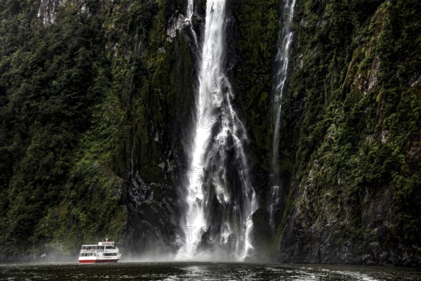 A mighty waterfall falls into the fjord, a boat glides past, surrounded by steep, green rocks, zero