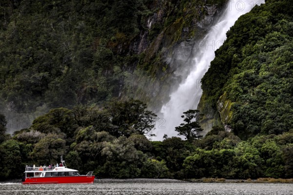 Boat sails past an impressive waterfall surrounded by lush vegetation, Milford Sound, New Zealand