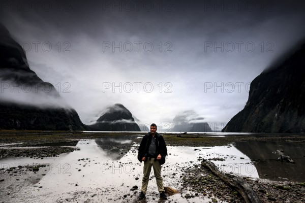 Man standing in front of a dramatic sky on the shore of a fjord with reflecting water, Milford Sound, New Zealand
