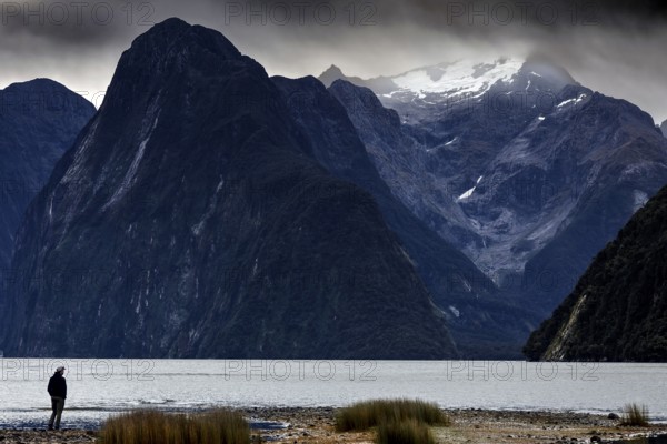 Lonely hiker on the banks of Milford Sound, with snow-capped mountains in the background, zero