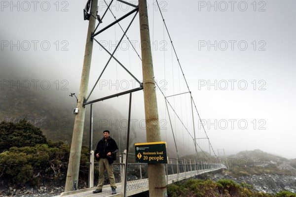 A man explores a high bridge in thick clouds of fog, Mt Cook, Hocker Valley Track, New Zealand