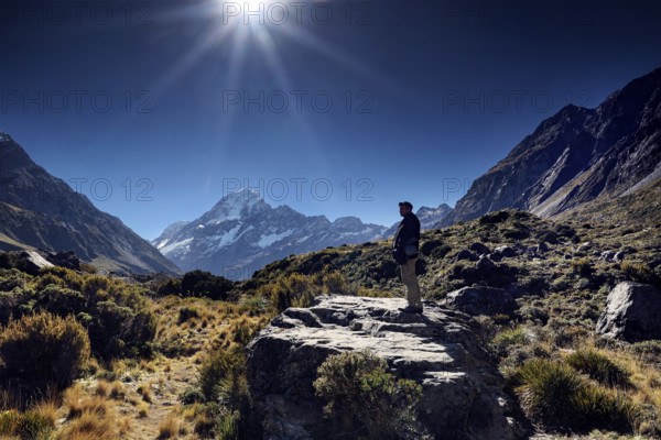 A hiker stands on a rocky trail with views of mountain peaks in sunshine, Mount Cook, New Zealand