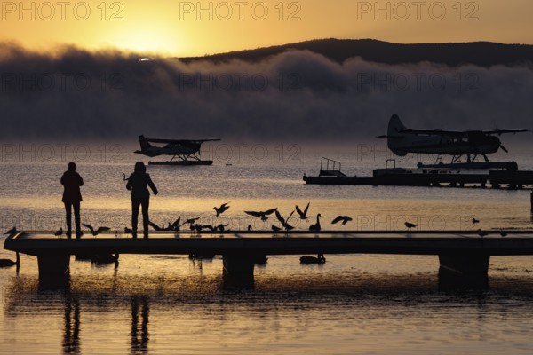 A seaplane on Lake Rotorua at sunrise, with silhouettes of people and birds on the dock, Rotorua, North Island, New Zealand