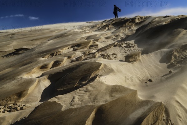 Person climbing large sand dunes during a sandstorm under a blue sky